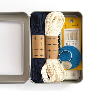May include: A metal tin containing two skeins of embroidery thread in navy blue and cream, each wrapped in a brown paper band with a black plus sign pattern. Includes a yellow package of gold-toned Sashiko needles, a blue circular thimble, and a white block.