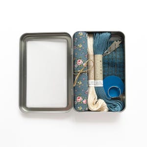 May include: An open, rectangular metal tin containing sewing supplies. Inside are spools of white and blue thread, a blue floral fabric, a blue fabric square, a blue thimble, needles, and a small silver tool. The tin is on a white background.