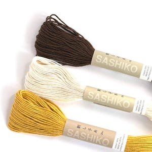 May include: Three spools of Sashiko thread in brown, cream, and mustard yellow. Each spool is wrapped with a paper label that includes the brand name and Japanese text. The thread is made of 100% cotton.