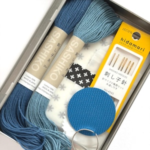 May include: A metal tin containing Sashiko embroidery supplies. The kit includes two spools of blue thread, needles, a blue fabric circle, a thimble, and patterned fabric. The text on the thread spools reads "Sashiko".