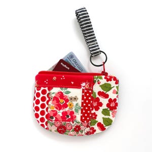 May include: A rectangular fabric coin purse featuring a patchwork design of red and white floral, polka dot, and solid patterns. It has a red zipper closure and a black and white striped wrist strap attached by a black metal ring. A card is inside.