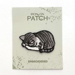 May include: An embroidered iron-on patch featuring a sleeping tabby cat with gray and white fur. The patch is displayed on a light green card with the text IRON-ON PATCH and EMBROIDERED printed in dark green lettering on the front of the card.