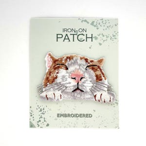 May include: An embroidered iron-on patch featuring a close-up of a ginger and white cat face with closed eyes and paws resting at the bottom. The patch is mounted on a light green card with the text IRON-ON PATCH and EMBROIDERED printed on it.