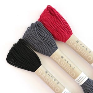 May include: Three skeins of Sashiko embroidery thread in black, dark gray, and red. Each skein is wrapped in a tan paper band labeled SASHIKO with Japanese characters and 20m. The thread is 100% cotton and made in Japan.