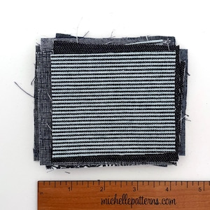 May include: A stack of square fabric swatches featuring various black and white patterns, including thin horizontal stripes and solid textures, positioned above a wooden ruler showing measurements in inches up to 5 inches.