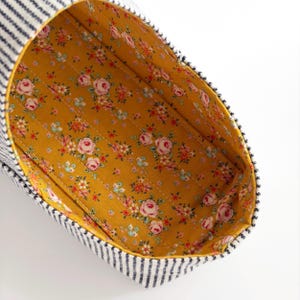 May include: A fabric pouch with a navy blue and white pinstriped exterior and a mustard yellow interior lining featuring a repeating floral pattern of pink roses and small green and white blossoms, showing visible horizontal quilted stitching.