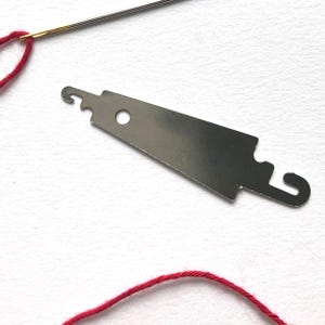 May include: A silver-colored needle threader with a hook and a hole, next to a needle with red thread. The threader is a flat, elongated tool with a curved end and a small circular hole.