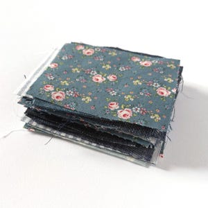 May include: A stack of square fabric swatches featuring a blue background with a repeating pattern of small pink roses and tiny white and yellow flowers. The edges of the fabric pieces are raw and frayed, showing a variety of dark blue and gray textures.