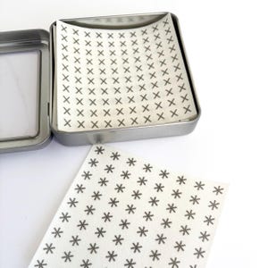 May include: An open, rectangular, silver metal tin with a white paper insert printed with small black crosses. A separate piece of paper with black star-shaped patterns lies next to the tin on a white surface.