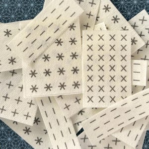 May include: An assortment of small, off-white rectangular and oblong fabric pieces. Each piece features a pattern of dark gray symbols, including asterisks, plus signs, and dashes. The pieces are scattered, creating a textured, layered effect.