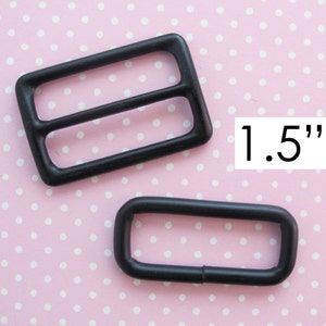 May include: Two black rectangular metal slide buckles, one with two bars and the other with one, are displayed against a pink polka dot background. The text "1.5" is visible in the image, indicating the size of the buckles.