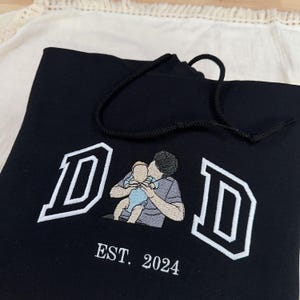May include: Black hoodie featuring white embroidered "DAD" lettering and an illustration of a parent holding a baby. The text "EST. 2024" is embroidered below. The hoodie has a black drawstring.
