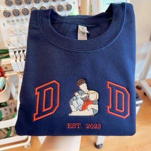 May include: A navy blue sweatshirt with a red embroidered design that says "DAD" with a family of three figures inside the letters. The text "EST. 2023" is embroidered below the design.