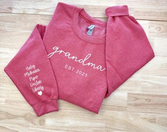 May include: A red sweatshirt with the text "grandma est. 2025" on the front. The left sleeve has the names "Haley, Mckenna, Piper, Declan, Liberty" printed on it.