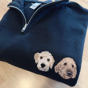 May include: Navy blue sweatshirt with a quarter-zip neckline. The sweatshirt features embroidered images of two golden doodle dog faces. The dogs have brown and tan fur, with black eyes and noses.