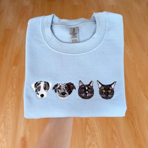 Peut inclure: Un sweat-shirt bleu clair avec des images brodées de deux chiens et deux chats. Les races de chiens sont un Border Collie et un croisé. Les chats sont noirs avec des yeux jaunes. Le sweat-shirt est plié.