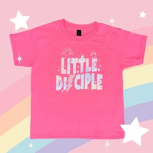 Könnte beinhalten: Ein leuchtend pinkfarbenes T-Shirt mit dem Schriftzug "LITTLE DISCIPLE" in einem farbenfrohen, im Batik-Stil gehaltenen Schriftzug. Das Design umfasst einen Regenbogen, ein Kreuz, eine Krone und einen Blitz. Das T-Shirt hat einen Rundhalsausschnitt.