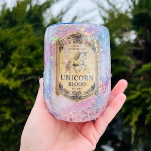 Puede incluir: Un vaso brillante con una etiqueta que dice "Mystic Apothecary Unicorn Blood No. 089". La taza es una mezcla de rosa, morado y azul con purpurina iridiscente. La etiqueta presenta una ilustración de unicornio.