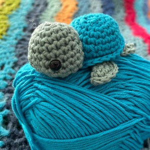 May include: A handcrafted crochet turtle in shades of blue and grey sits on a ball of turquoise yarn. The turtle features a grey head with a black button eye, a blue shell, and grey flippers. The background is a colourful, patterned blanket.