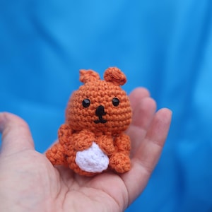May include: A handmade orange crochet squirrel with black eyes and a white belly. The small stuffed animal is held in a hand against a blue background. The squirrel has a cute, cartoonish design.
