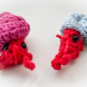 May include: Two handmade crochet jellyfish toys. One has a pink cap, the other a light blue cap. Both have red bodies, black eyes, and small tentacles. These whimsical toys are perfect for children.