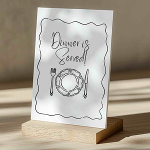 May include: A white sign with black text and a simple illustration of a plate, fork, and knife. The text reads "Dinner is Served!"