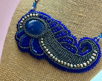 Puede incluir: Un collar artesanal con una piedra grande, ovalada y azul oscuro en el centro. El collar presenta un intrincado trabajo de abalorios en tonos azules y plateados, creando un diseño detallado y llamativo. El collar se exhibe sobre una forma cubierta de arpillera.