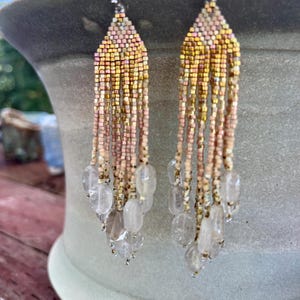 May include: Beaded earrings with a fringe design. The earrings combine pink, gold, and clear beads. The top section has a solid pattern, leading to a fringe of clear, oval-shaped beads. A stylish accessory.