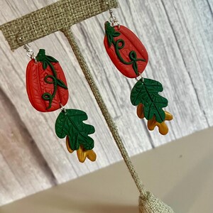 May include: A pair of fall-themed earrings. Each earring features a red pumpkin with green vines, a green leaf, and a gold leaf. The earrings are displayed on a tan earring stand against a light wood background.