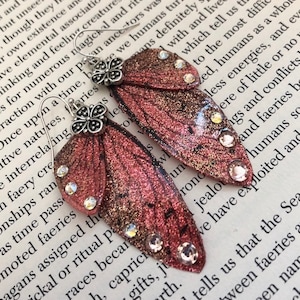 Puede incluir: Pendientes hechos a mano con un diseño de ala de mariposa. Los pendientes tienen un efecto de purpurina rosa rojizo y dorado, adornados con pedrería redonda y transparente. La parte superior de los pendientes tiene un acento de mariposa plateada.