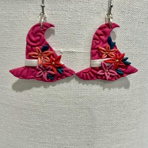May include: A pair of pink witch hat earrings. The hats are decorated with a white band and colorful flowers. The earrings are made of a textured material and have silver-colored hooks.