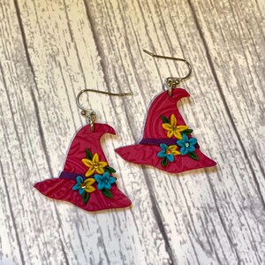 May include: A pair of witch hat earrings. The earrings are a vibrant pink with a textured pattern. Each hat is adorned with yellow and blue flowers, a purple band, and silver-colored ear hooks. The earrings are a whimsical accessory.