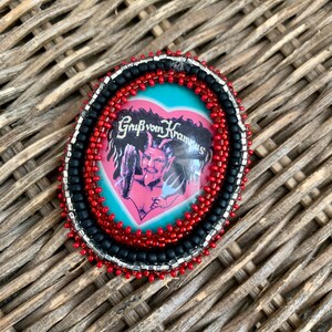 May include: Oval-shaped pin with a beaded border. The pin features a central image of a devil-like figure within a pink heart, set against a light blue background. The text "Grüß vom Krampus" is visible. The border is composed of red, black, and silver beads.