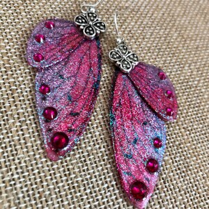 Pode incluir: Um par de brincos de asas de borboleta feitos à mão. As asas são em tons de rosa e roxo com glitter e pequenas pedras preciosas vermelhas redondas. Os brincos têm tops decorativos prateados e ganchos prateados.