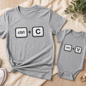 May include: A gray t-shirt with the text "ctrl + C" and a gray baby onesie with the text "ctrl + V". The clothing items are on a cream-colored blanket.