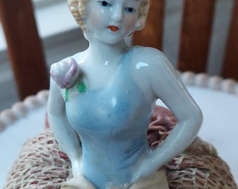 May include: A porcelain figurine of a woman with blonde hair and a light blue dress. A pink rose is pinned to the dress. The figurine is seated on a pink and beige fabric base, with a bow at the waist. The figurine is a vintage collectible.