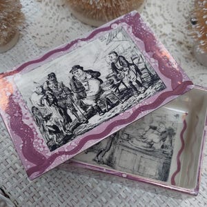 May include: A rectangular ceramic trinket box with a lid. The lid features a black and white illustration of people in a market scene, framed by a wavy, pink border. The box interior is white with a matching illustration.