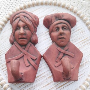 May include: Two terracotta wall hooks shaped like people's heads. The hooks are painted in a reddish-brown color and have a rustic look. Each hook has a simple, rounded face and a small, round hook protruding from the bottom.