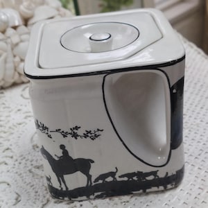 May include: White ceramic creamer with a square shape and a lid. The creamer features a black silhouette design of a person on a horse, dogs, and foliage. The handle and rim are outlined in black.