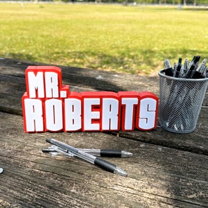 May include: Red and white plastic sign with the text "MR. ROBERTS" on a wooden surface with black pens in a wire mesh cup.