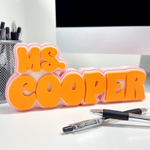 May include: A personalized desk nameplate with the text "MS. COOPER" in orange bubble letters, outlined in white and pink. Two pens are in the foreground. A metal pen holder is in the background.