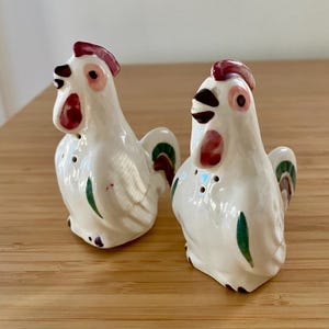 May include: A pair of white ceramic salt and pepper shakers shaped like roosters. Each figure features a red comb and wattle, black eyes and beak, and green accents on the wings and tail feathers. The shakers have small holes on the chest area.