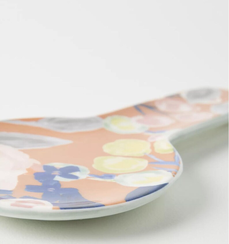 May include: A ceramic spoon rest with a peach-colored background and a floral pattern in blue, yellow, and pink. The spoon rest has a curved shape and a white rim, perfect for the kitchen.