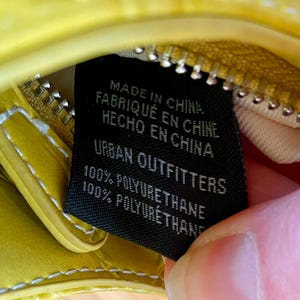 Puede incluir: Primer plano de un bolso amarillo con cremallera y etiqueta negra. La etiqueta dice "MADE IN CHINA, FABRIQU&Eacute; EN CHINE, HECHO EN CHINA, URBAN OUTFITTERS, 100% POLYURETHANE". El bolso es de material brillante.
