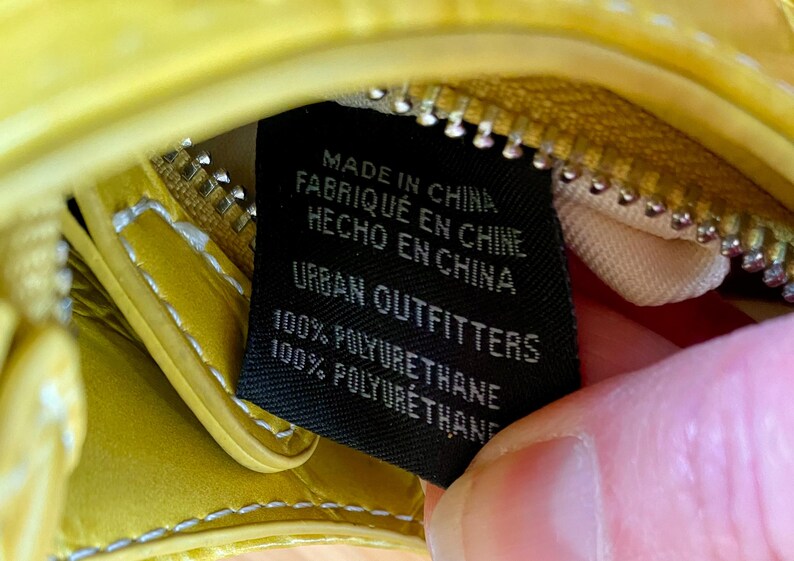Puede incluir: Primer plano de un bolso amarillo con cremallera y etiqueta negra. La etiqueta dice "MADE IN CHINA, FABRIQU&Eacute; EN CHINE, HECHO EN CHINA, URBAN OUTFITTERS, 100% POLYURETHANE". El bolso es de material brillante.