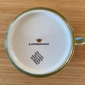 Puede incluir: Una taza de cer&aacute;mica verde con interior blanco, con el texto "by ANTHROPOLOGIE" y un gr&aacute;fico de abeja. La base de la taza tambi&eacute;n incluye texto que indica que es solo para lavar a mano y est&aacute; hecha en China.