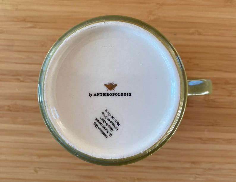 Puede incluir: Una taza de cer&aacute;mica verde con interior blanco, con el texto "by ANTHROPOLOGIE" y un gr&aacute;fico de abeja. La base de la taza tambi&eacute;n incluye texto que indica que es solo para lavar a mano y est&aacute; hecha en China.