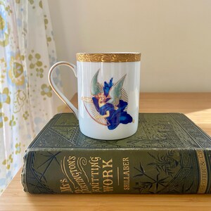 May include: A white ceramic mug with a gold rim and handle, decorated with an angel in blue robes and wings. The mug sits on a vintage green book with gold lettering. The book is titled "Mrs. Barrington's Knitting Work".
