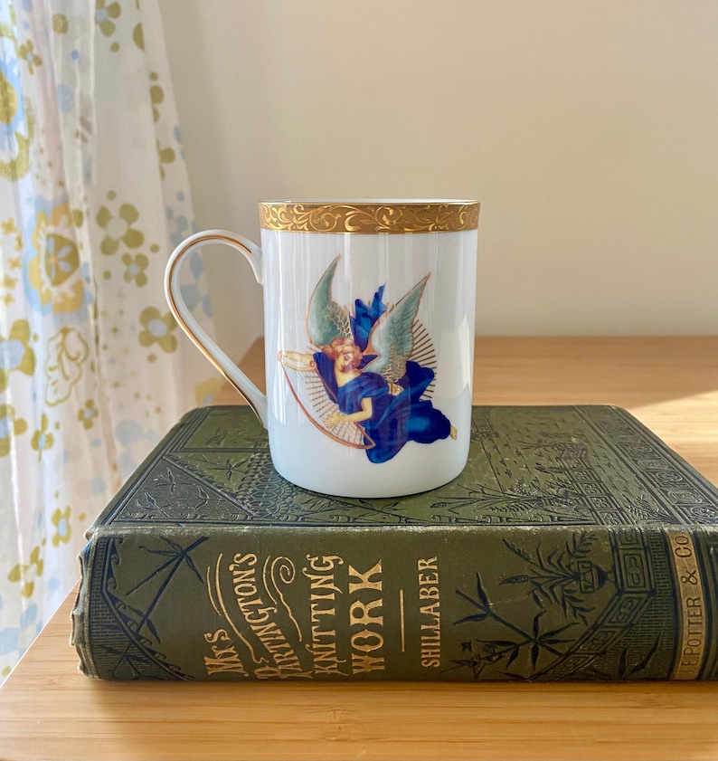 May include: A white ceramic mug with a gold rim and handle, decorated with an angel in blue robes and wings. The mug sits on a vintage green book with gold lettering. The book is titled "Mrs. Barrington's Knitting Work".