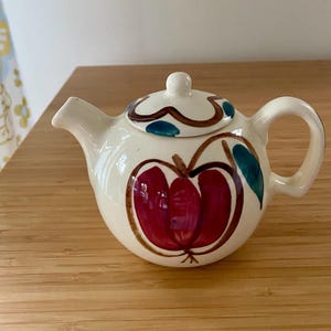 May include: A cream-colored ceramic teapot featuring a hand-painted red apple design with green leaves. The teapot has a matching lid with a small knob handle, a curved spout, and a rounded handle. The painted artwork includes brown outlines.